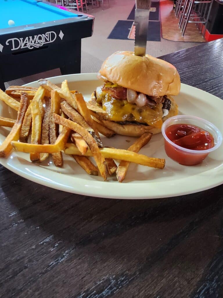 Juicy blue cheese bacon burger with grilled onions, hand-cut fries, and ketchup served at Jimmy’s Pro Billiards near a Diamond pool table
